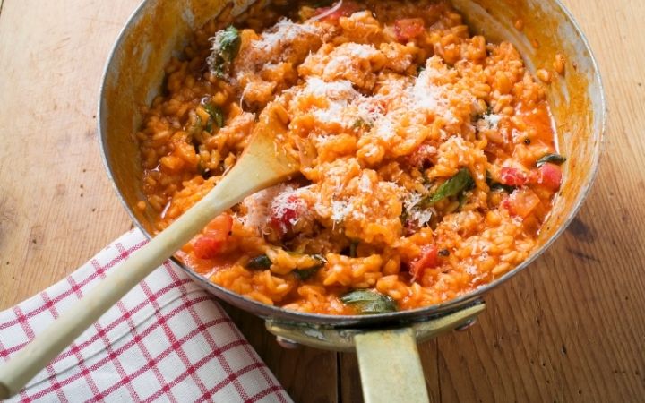 Risotto in a pot with wooden spoon on a table whit a cloth napkin beside - The Proud Italian